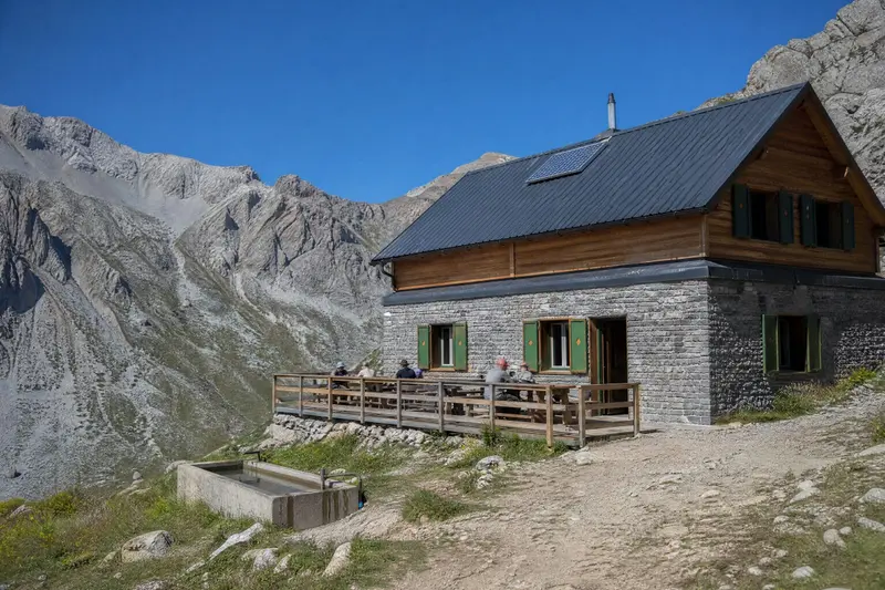 Refuge de montagne en pierre et en bois, aux volets verts, avec une terrasse où des randonneurs se reposent au pied de crêtes rocheuses.