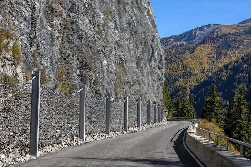 Route de montagne au pied d'une falaise couverte de filets métalliques et protégée par une rangée de barrières pare-blocs.